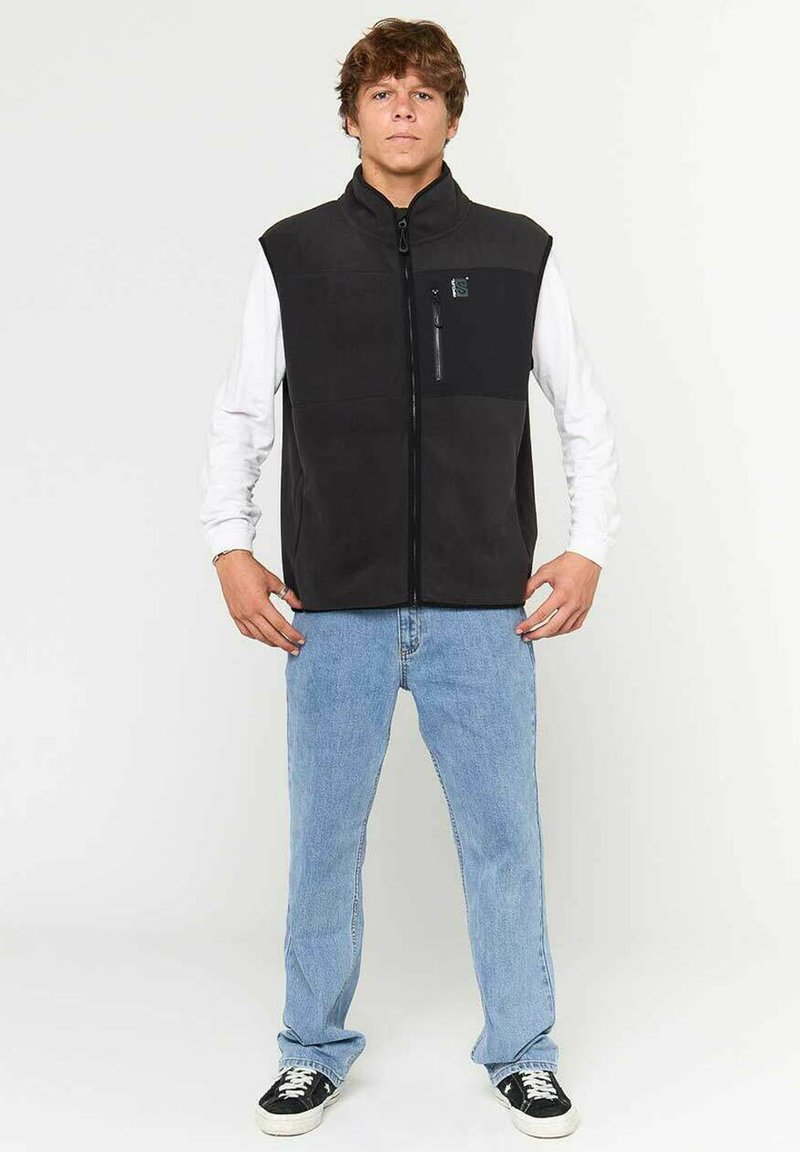 Chaleco de forro polar negro con cuello alto, cierre frontal de cremallera y un bolsillo con cremallera en el pecho izquierdo. Lleva puesto una camisa blanca de manga larga y jeans azul.