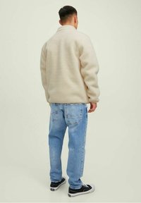 Uomo che indossa una giacca di pile beige, jeans azzurri e sneakers nere, in piedi rivolto di spalle su uno sfondo chiaro e uniforme.