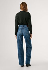Jean haut à taille haute bleus avec une jambe droite, présentant un motif subtil et des poches arrière, associé à un haut à manches longues foncé.