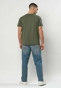 Olivengrønn t-skjorte med korte ermer og avslappet passform, vist sammen med lyse blå jeans og hvite joggesko, sett bakfra.