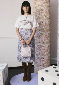 Weißes Grafik-T-Shirt mit floralem Design, kombiniert mit einem gemusterten lavendelfarbenen Rock, braunen knielangen Stiefeln und einer helllila Tasche mit Tierillustrationen.