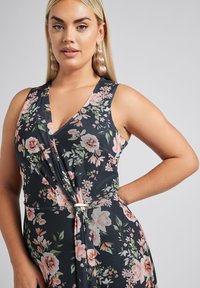 Kukkakirjallinen kietaisumekko tummansinisellä pohjalla, jossa on vaaleanpunaisia ja vihreitä kukkakuvioita. V-kaula-aukko, sidottu vyötärö ja kultainen varustelu.