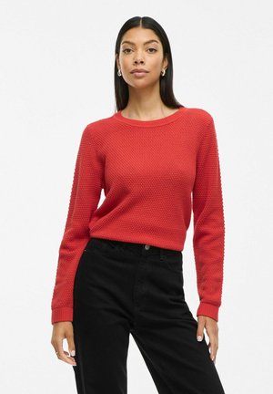 Rode gebreide trui met textuurpatroon, ronde halslijn en geribbelde manchetten; gecombineerd met zwarte high-waisted broek.