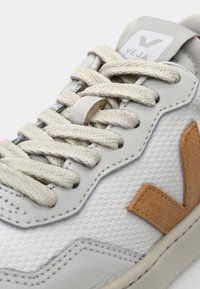 Sneaker with a textured white mesh upper, beige laces, and a prominent brown V logo on the side. Gray heel tab features brand name.