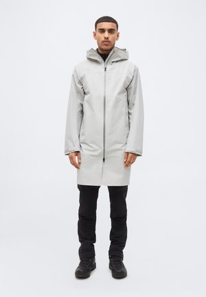 Férfi világosszürke kapucnis esőkabátban, felhúzott cipzárral, fekete nadrágban és fekete csizmában, egyszerű fehér háttér előtt állva.