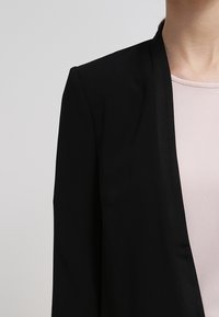 Osoba nosząca czarną marynarkę na jasnoróżowej, teksturowanej bluzce, przedstawiona od szyi do połowy torsu na neutralnym tle.