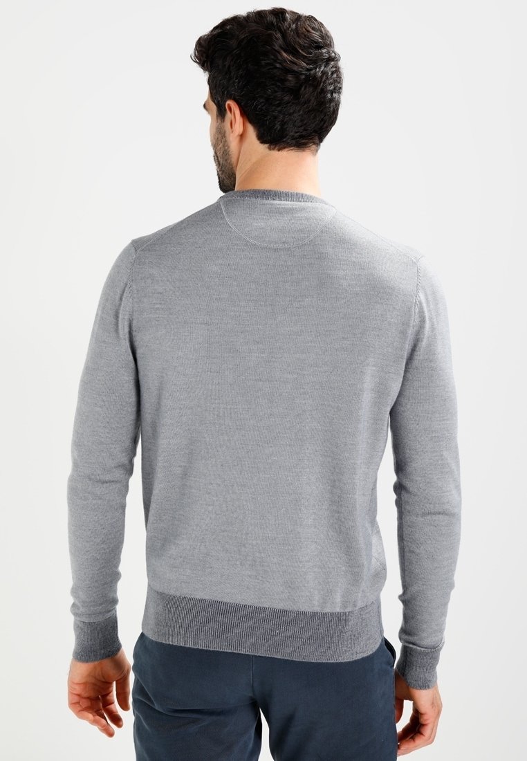 Homme aux cheveux bouclés foncés portant un pull gris clair à manches longues et un pantalon sombre, vu de dos sur un fond clair.