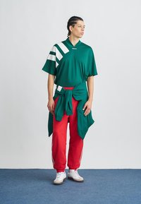 Groen shirt met korte mouwen en witte strepen, rode broek en een groene trui om de taille geknoopt. Witte sneakers maken de look af.