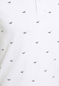 Tissu blanc avec de petits logos d'oiseaux noirs uniformément espacés et une vue partielle d'un col blanc à boutons.