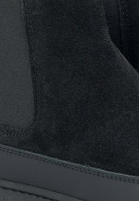 Black suede ankle boot featuring elastic side panels and a flat rubber sole. Detailed stitching along the sole edge. Matte finish.