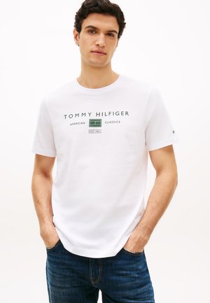 Junger Mann trägt ein weißes Tommy Hilfiger T-Shirt mit grünem Text und Grafik, Hände in den Taschen, steht vor einem schlichten hellen Hintergrund.