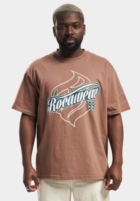 Brown cotton t-shirt featuring a large printed Rocawear logo in white and teal, short sleeves, and a relaxed fit.