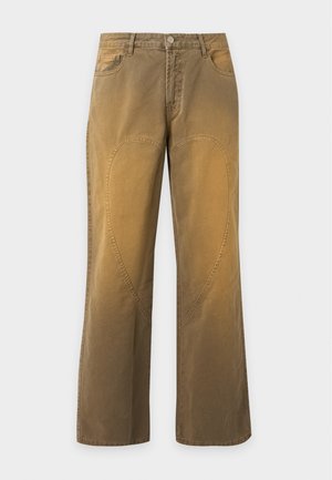 Homme debout portant un t-shirt noir uni à manches courtes, un pantalon marron ample avec des coutures courbes et des baskets noires sur un fond gris clair.