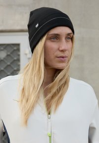 Black knit beanie with a ribbed texture and a small logo. Model wears a white zip-up top with green accents and blue detail along the sides.