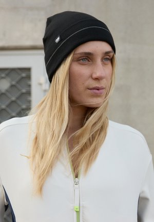 Bonnet en tricot noir avec une texture côtelée et un petit logo. Le modèle porte un haut zippé blanc avec des accents verts et un détail bleu sur les côtés.