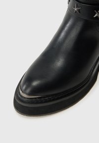 Bota de couro preta com bico arredondado, sola espessa e tiras com tachinhas metálicas em formato de estrela sobre um fundo branco.