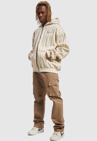 Kremfarget zip-hettegenser med bølgete svart mønster, sammen med khaki cargo-bukser med flere lommer og hvite joggesko.