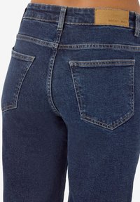 Tmavě modré džíny z denimu s hladkou texturou, s zadními kapsami a koženou hnědou štítkem s nápisem "NOISY MAY" na pásku.