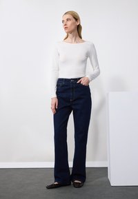 Weißer langärmeliger Body, kombiniert mit dunkelblauen Schlagjeans. Das Outfit zeichnet sich durch eine taillierte Silhouette mit klassischer Denim-Textur und hoher Taille aus.