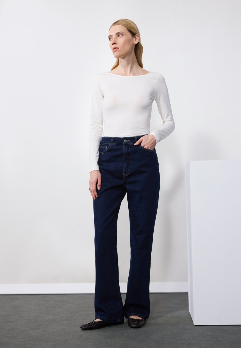 Weißer langärmeliger Body, kombiniert mit dunkelblauen Schlagjeans. Das Outfit zeichnet sich durch eine taillierte Silhouette mit klassischer Denim-Textur und hoher Taille aus.