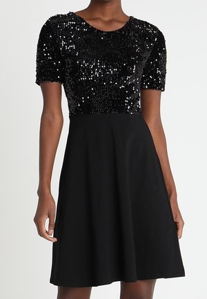 Schwarzes Kleid mit kurzen Ärmeln, paillettenbesetztem, tailliertem Mieder und schlichter A-Linien-Rock, getragen von einer stehenden Person vor einfarbigem Hintergrund.