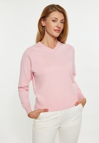 Helles rosa Pullover mit V-Ausschnitt, gerippten Bündchen und einem glatten, strukturierten Stoff. Das Model trägt weiße Hose und hat die Hände in den Taschen.