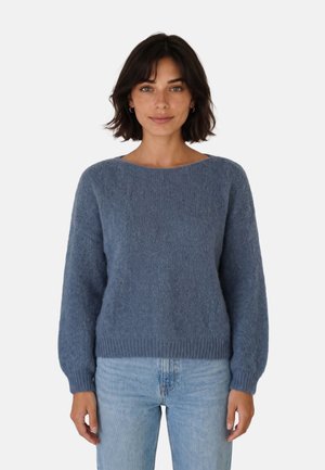 Mujer con cabello oscuro hasta los hombros, que lleva un jersey azul con textura y vaqueros azul claro, de pie frente a un fondo liso y claro.