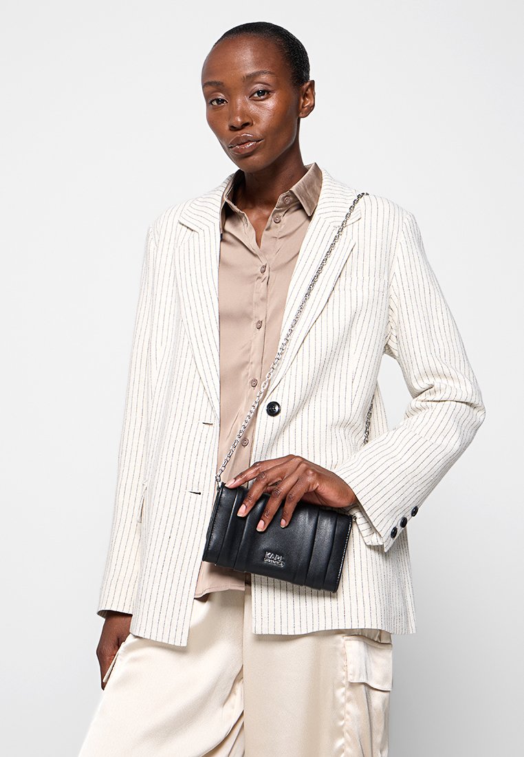 Sac en cuir noir en bandoulière avec une texture matelassée, doté d'une bandoulière en chaîne argentée. Le modèle porte un blazer beige à rayures et une chemise assortie.