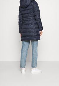 Abrigo acolchado azul marino con capucha, diseño acolchado horizontal y mangas largas, combinado con jeans azul claro y zapatillas blancas.