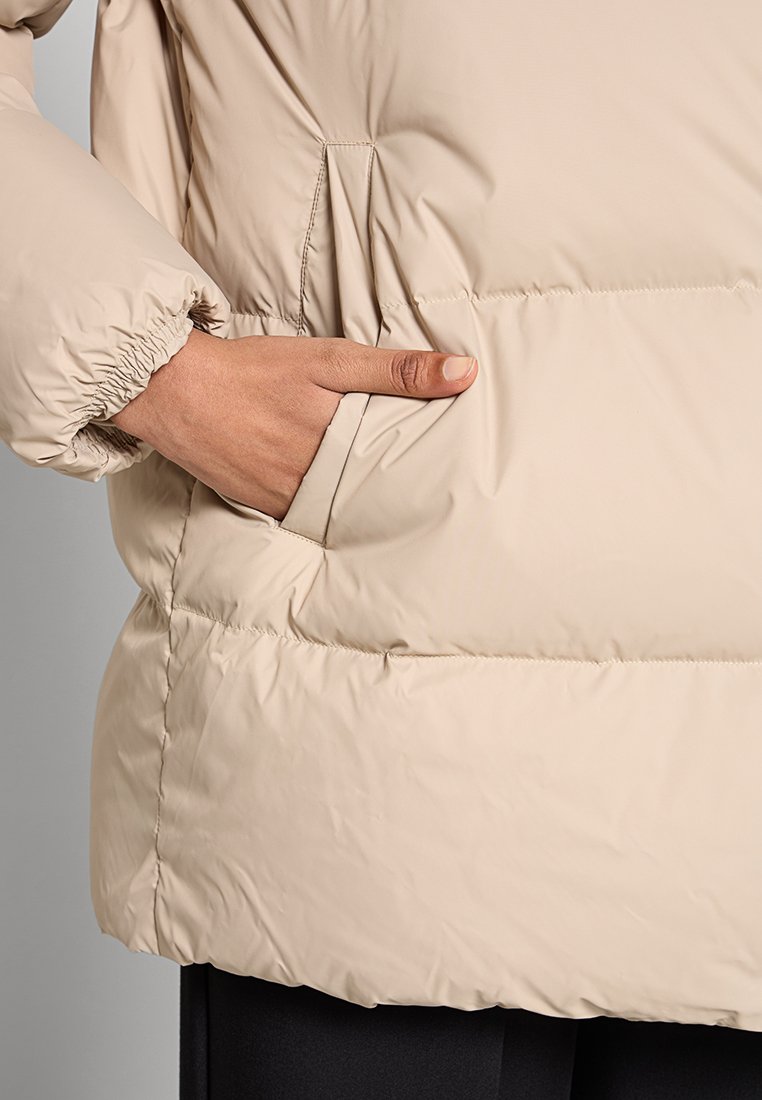 Beżowa kurtka puchowa o miękkim, błyszczącym wykończeniu. Posiada elastyczne mankiety i boczną kieszeń, prezentując pikowany wzór dla lepszej izolacji.
