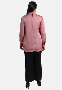 Blusa di raso rosa chiaro con maniche lunghe, collo alto e chiusura con bottoni posteriori. Indossata con pantaloni neri a gamba larga e tacchi.