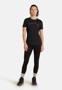 T-shirt nero a maniche corte realizzato in un tessuto morbido, con un design marrone sul petto. Abbinato a leggings neri e scarponi da trekking neri.