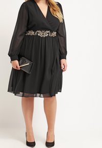Czarna sukienka wrap z długimi przezroczystymi rękawami, ozdobnym paskiem w talii i zwiewnym dołem. Połączona z czarną kopertówką i wysokimi obcasami.