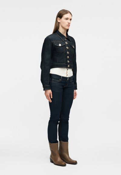 Γυναίκα που φοράει σκούρο τζιν μπουφάν και τζιν παντελόνι, λευκό τοπ με κουμπιά και καφέ μπότες μέχρι τη μέση της γάμπας, σε λευκό φόντο.