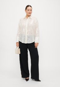 Sequined white shirt with a collar, button front, and cuffed sleeves, paired with wide black trousers and a cream handbag.