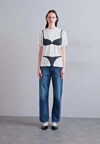 White T-shirt featuring black bra and panty print; paired with blue straight-leg jeans and black loafers with bows. Simple, casual design.