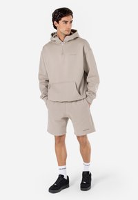 Beige zip-up hoodie med frontlomme, parret med beige shorts. Har blødt stof, afslappet pasform og sorte sneakers med hvide sokker.