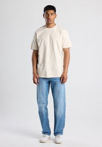 Vaaleanbeige lyhythihainen t-paita pehmeästä puuvillasta, yhdistettynä vaaleansinisiin suoriin farkkuihin ja valkoisiin lenkkareihin. Yksinkertainen, minimalistikko muotoilu.