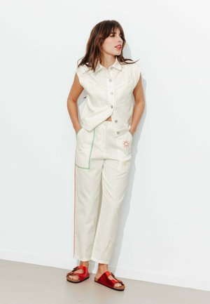 Femme en haut blanc sans manches boutonné et pantalon blanc brodé, debout les mains dans les poches, portant des sandales rouges devant un mur blanc.