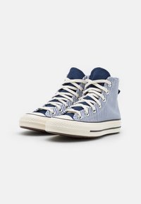 Høy-top sneakers med marineblå og hvite striper i lerret, kremfarget snøring, gummisåle og svart akse stripe langs bunnen.