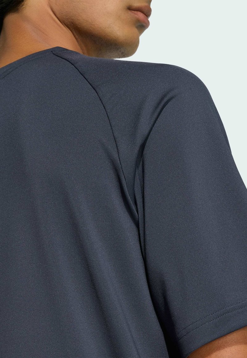 Primer plano de una persona que lleva una camiseta de manga corta gris oscuro, mostrando el hombro, el cuello y parte del rostro contra un fondo claro.