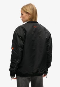 Sort bomberjakke med en glat, blank overflade. Har et orange bølgegrafik på ryggen og en lynlåslomme på ærmet. Elastiske manchetter og kant.