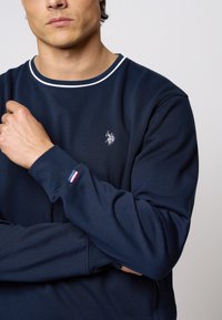 Tamsiai mėlynas džemperis su balta apykakle ir išsiuvinėtu logotipu krūtinėje. Rankovėje yra mažas trijų spalvų akcentas raudonai, baltai ir mėlynai.