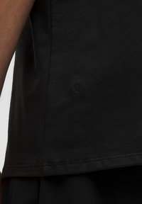 Svart t-shirt gjord av mjukt tyg. Har en rundad nederkant och en liten, präglad logotyp på den nedre sidan. Slät textur och minimalistisk design.