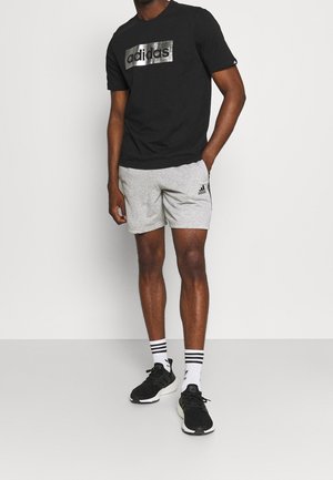 Hombre vistiendo una camiseta negra de Adidas, pantalones cortos grises de Adidas, zapatillas negras y calcetines blancos de Adidas, de pie con una mano en el bolsillo.