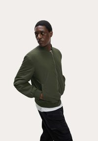 Giacca bomber verde oliva con zip frontale, polsini a coste e due tasche laterali, indossata sopra una camicia bianca, esposta su uno sfondo chiaro.