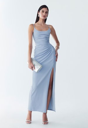 Femme portant une robe sans manches bleu clair froncée avec une fente à la cuisse, tenant une pochette argentée et portant des sandales et des bijoux argentés sur fond blanc.
