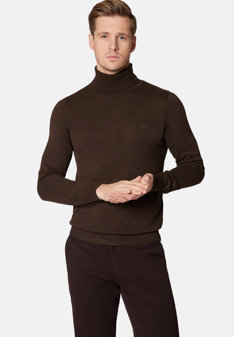 Brązowy sweter typu golf z dopasowanym krojem, z prążkowanymi mankietami i dołem, z małym logo na piersi, noszony z ciemnymi spodniami.