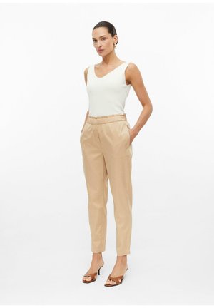 Femme debout avec les mains dans les poches, portant un top blanc sans manches, un pantalon beige taille haute et des sandales marron à talons sur fond blanc.