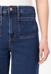Vaqueros de mezclilla azul oscuro con una cintura alta, cinco bolsillos y detalles de costura en color naranja contrastante. Textura suave con un material duradero.